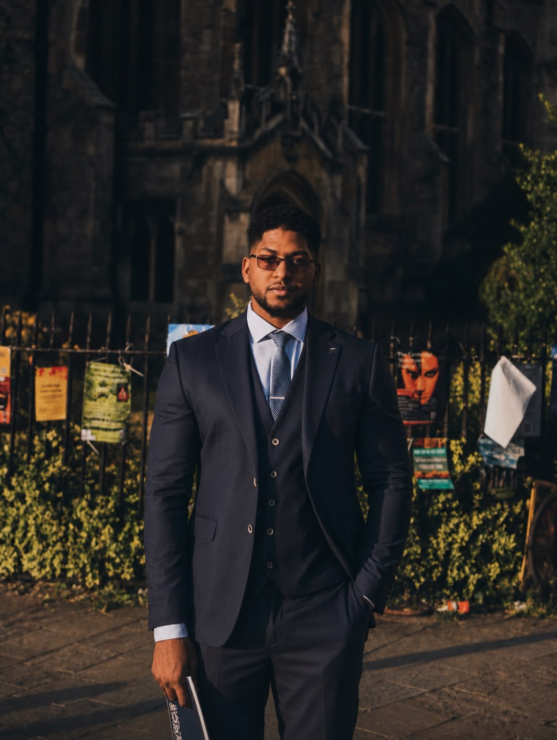 Moayed Musa — Cambridge, UK. Editorial portrait in navy three-piece suit outside a sunlit gothic stone church with a book. Software engineer and creative developer.